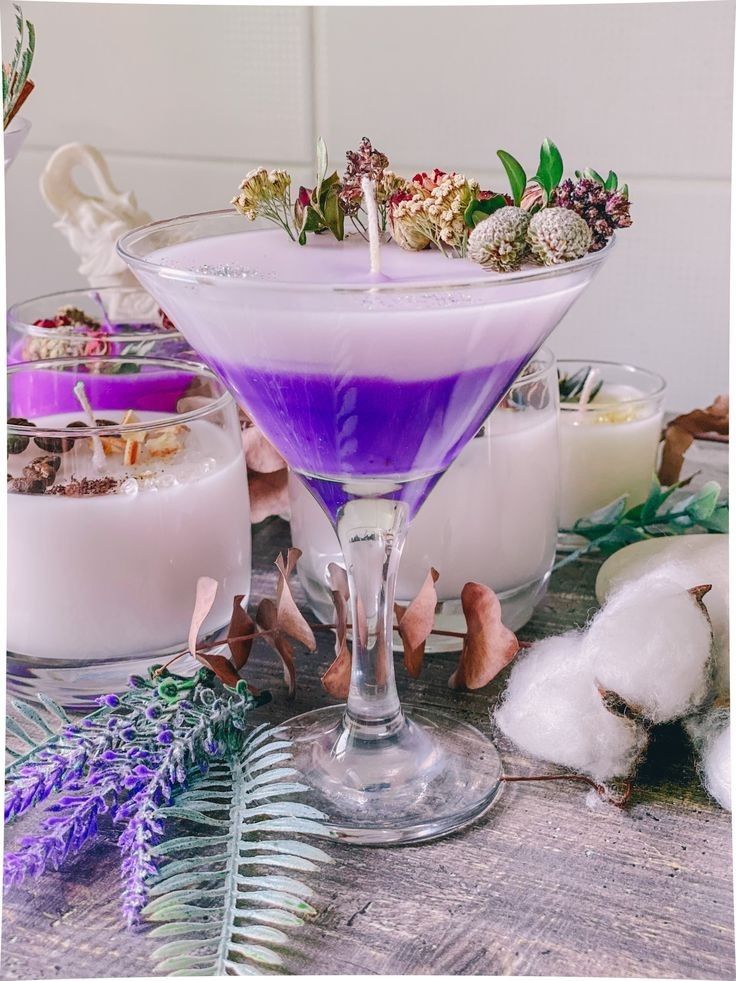 Purple cocktail in a martini glass with floral garnish on a table with candles and plants.