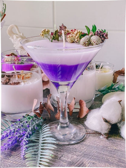Purple cocktail in a martini glass with floral garnish on a table with candles and plants.